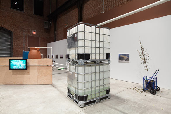 Tue Greenfort, vue de l’exposition / view of the exhibition « Garbage Bay », Sculpture Center, Long Island City, 2013. Photo : Jason Mandella. À partir de l’écosystème de Jamaica Bay, Greenfort a créé une installation avec un arbre provenant d’une campagne de reboisement menée par le New York Restoration projet (I’m One in a Million, 2013), un impluvium (SculptureCenter Impluvium – Ecobulk, 2013) relié à une sculpture filtrante triangulaire (The Great Gateway, 2013) où l’eau ruisselle parmi paillettes et tubulures fluo. / Based on the ecosystem of Jamaica Bay, Greenfort has created an installation with a tree coming from a re-afforestation campaign undertaken by the New York Restoration projet (I’m One in a Million, 2013), and an impluvium (Sculpture Center Impluvium – Ecobulk, 2013) connected to a triangular filtering sculpture (The Great Gateway, 2013) where water flows among spangles and fluorescent pipes.